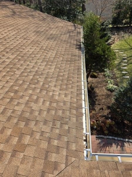 A high-angle view of a brown shingled roof with a white gutter running along the edge, overlooking a garden area.