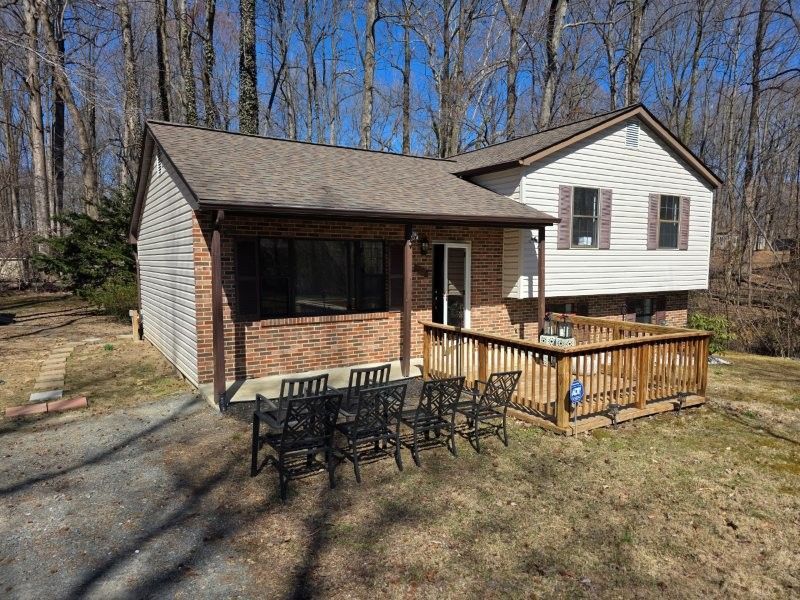 A split-level house with a brick facade and a wooden deck featuring patio chairs in a wooded setting under a blue sky.