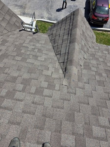 A view from a roof showing grey shingles, a valley, a metal ladder, and a car parked on the asphalt below.