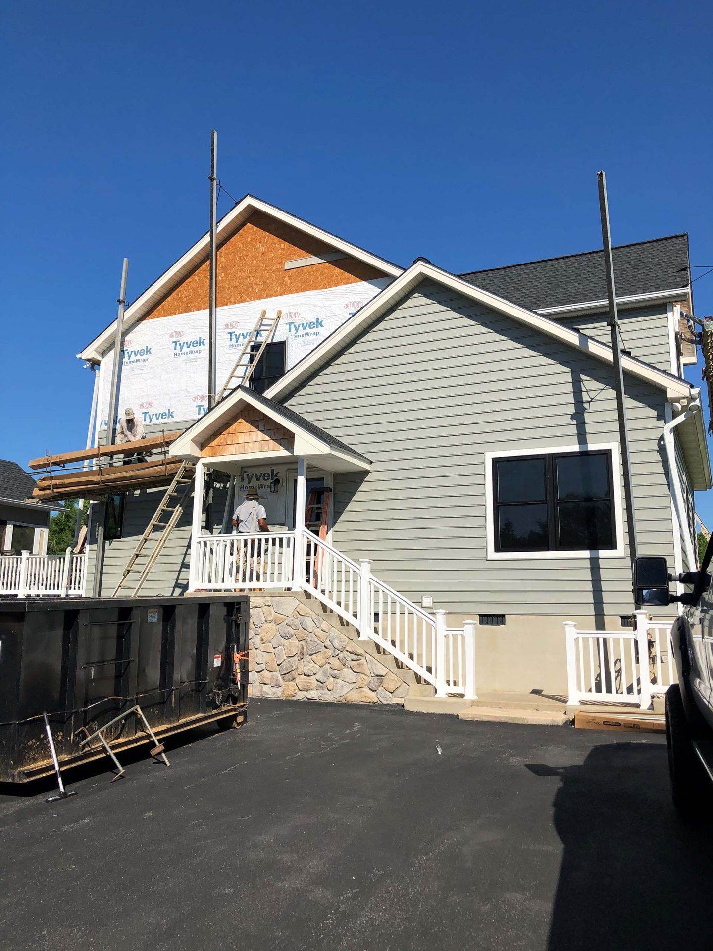 A partially sided two-story house under construction with scaffolding, a stone staircase, and a large dumpster in front.